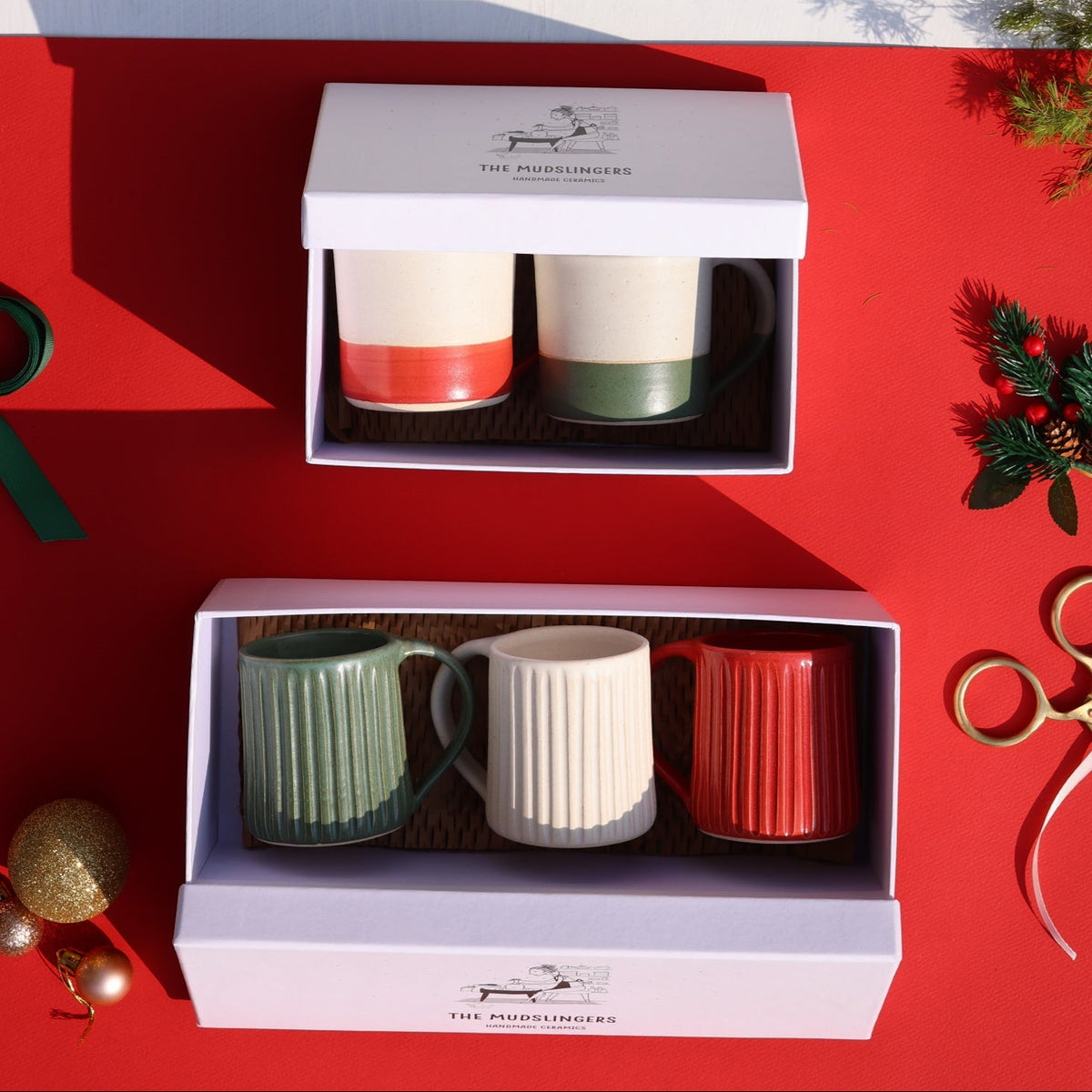 Two boxed holiday mug sets arranged on a split white and red surface with Christmas decorations. The top set contains two cream mugs with orange and green bands.