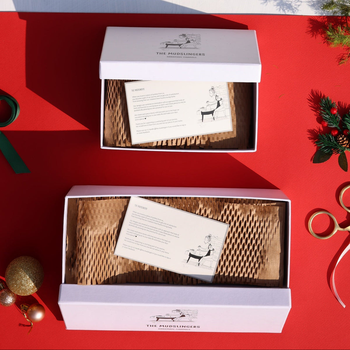 Two white gift boxes with lids are arranged on a split red and white background with holiday decorations. The boxes are open, showing brown packing material and a printed card.