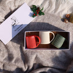 A white gift box with a red ribbon bow and the printed brand name 'THE MUDSLINGERS' is held by hands wearing a red sweater, set against a cozy background with ceramic mugs, a green knit blanket, and holiday decorations.