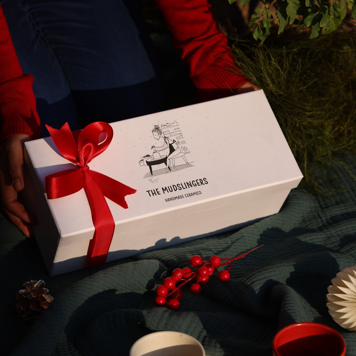 A white gift box with a red ribbon bow and the printed brand name 'THE MUDSLINGERS' sits on a dark green blanket, surrounded by ceramic mugs and holiday decorations. A person in a red sweater is partially visible holding the box.