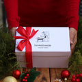 A person in a red sweater holds a white gift box tied with a red ribbon. The box is printed with the brand name 'THE MUDSLINGERS' and a small black line drawing.