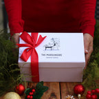 A person in a red sweater holds a white gift box tied with a red ribbon. The box is printed with the brand name 'THE MUDSLINGERS' and a small black line drawing.