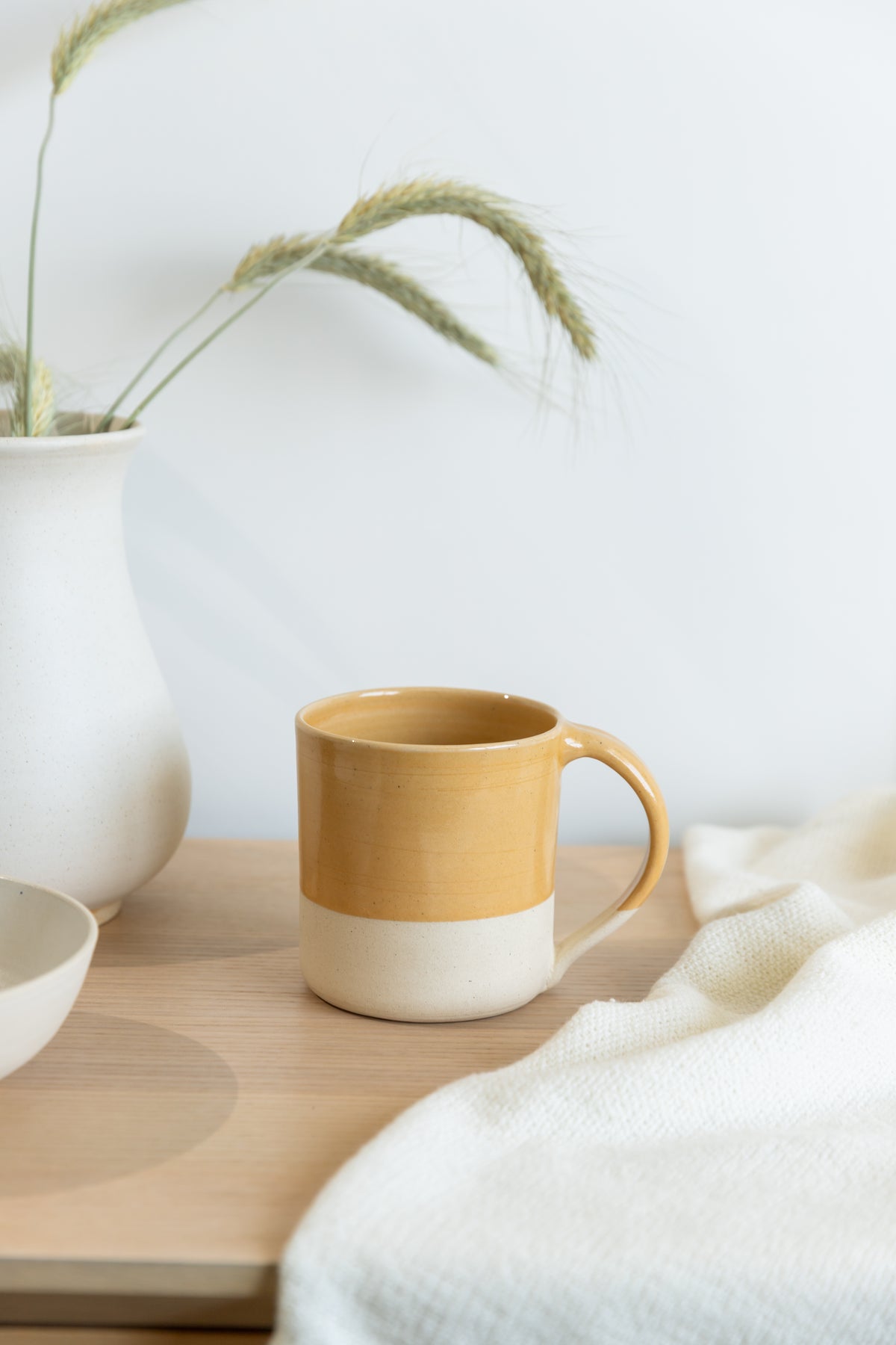 Brown and white ceramic mug on a wooden surface with a white vase and plant in the background.