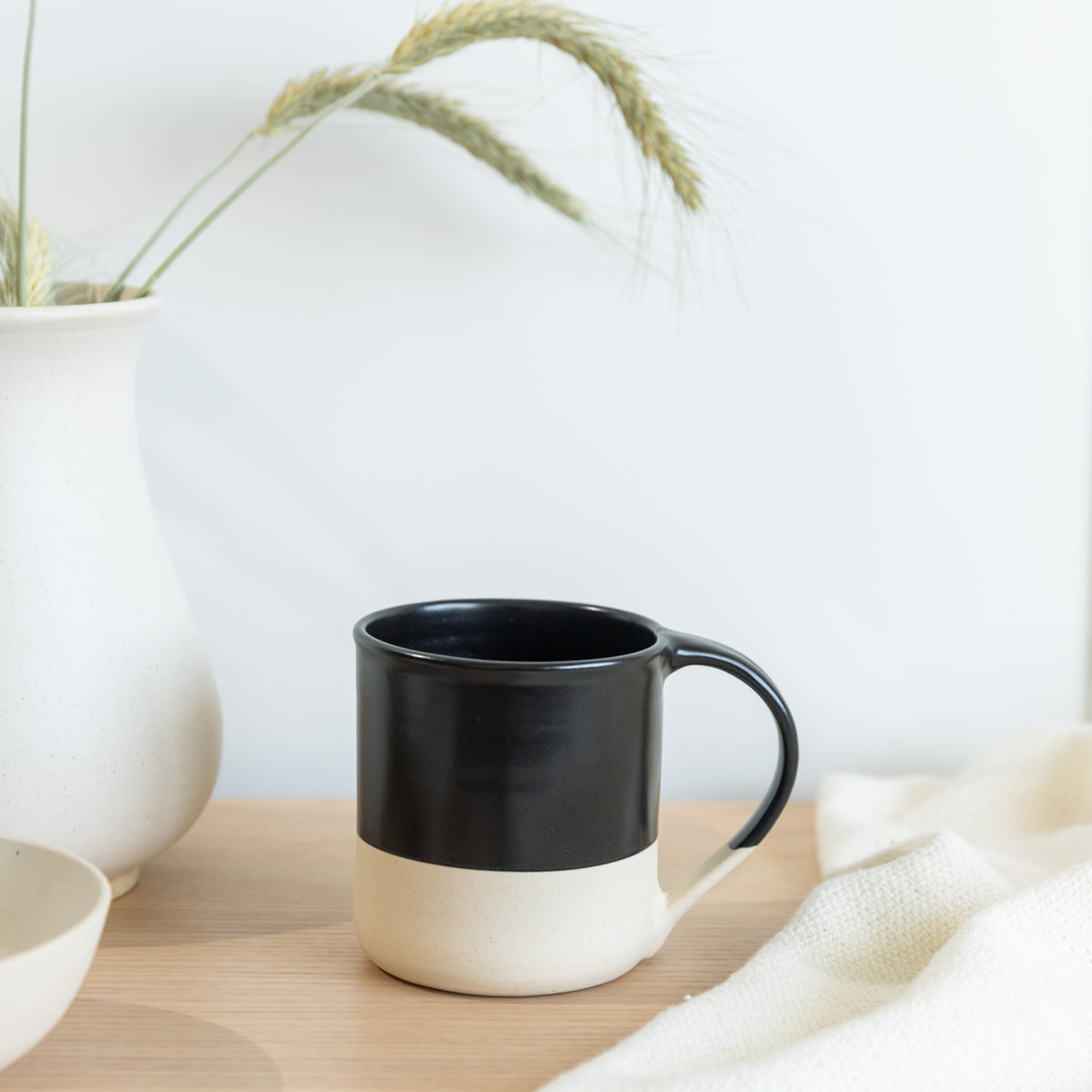 A ceramic mug with a two-tone design, featuring a glossy black upper half and a matte cream lower half, resting on a light wooden surface next to a white vase with dried wheat.