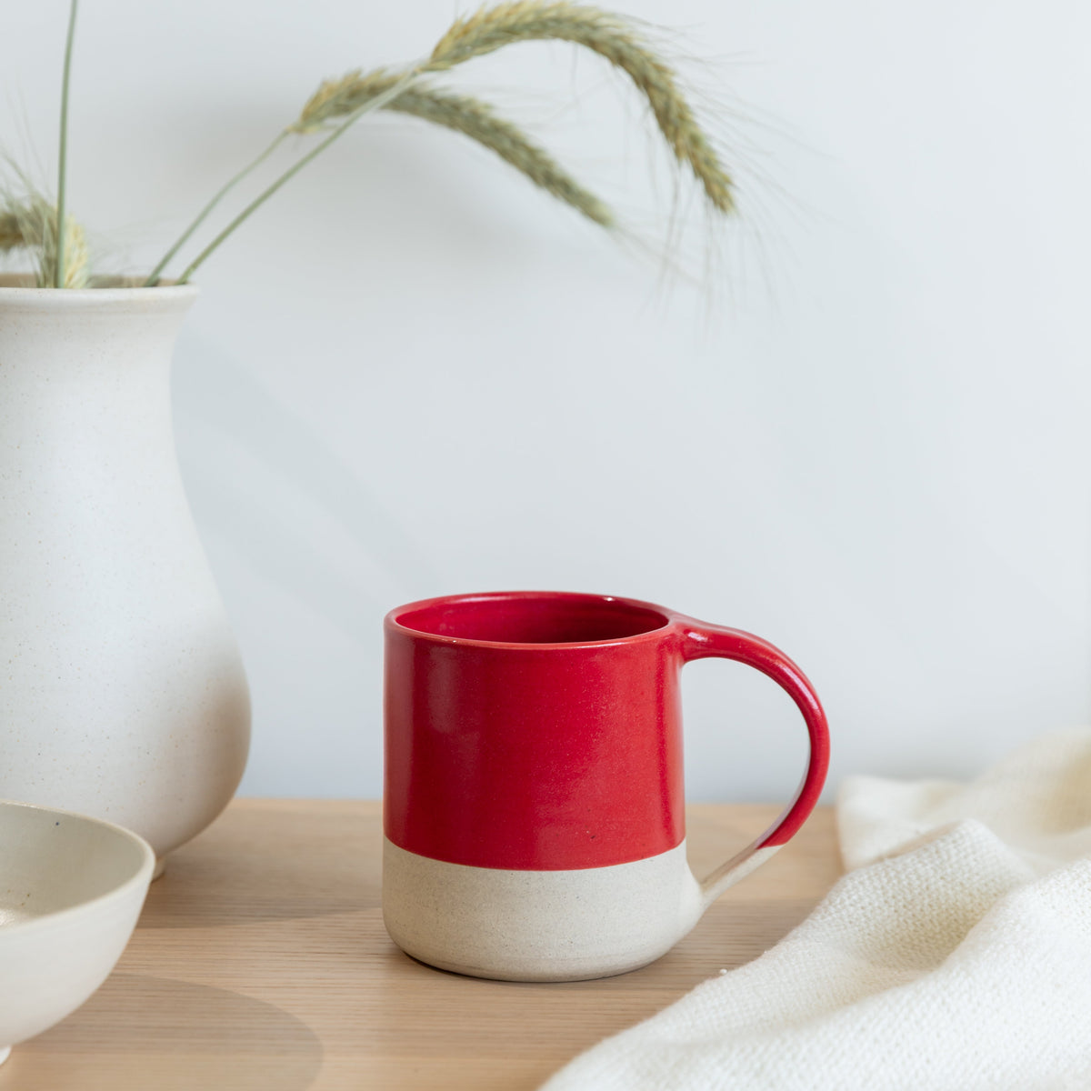 A ceramic mug with a two-tone red and cream lower half, placed on a light wooden table next to a white vase with dried wheat and a white textured cloth.